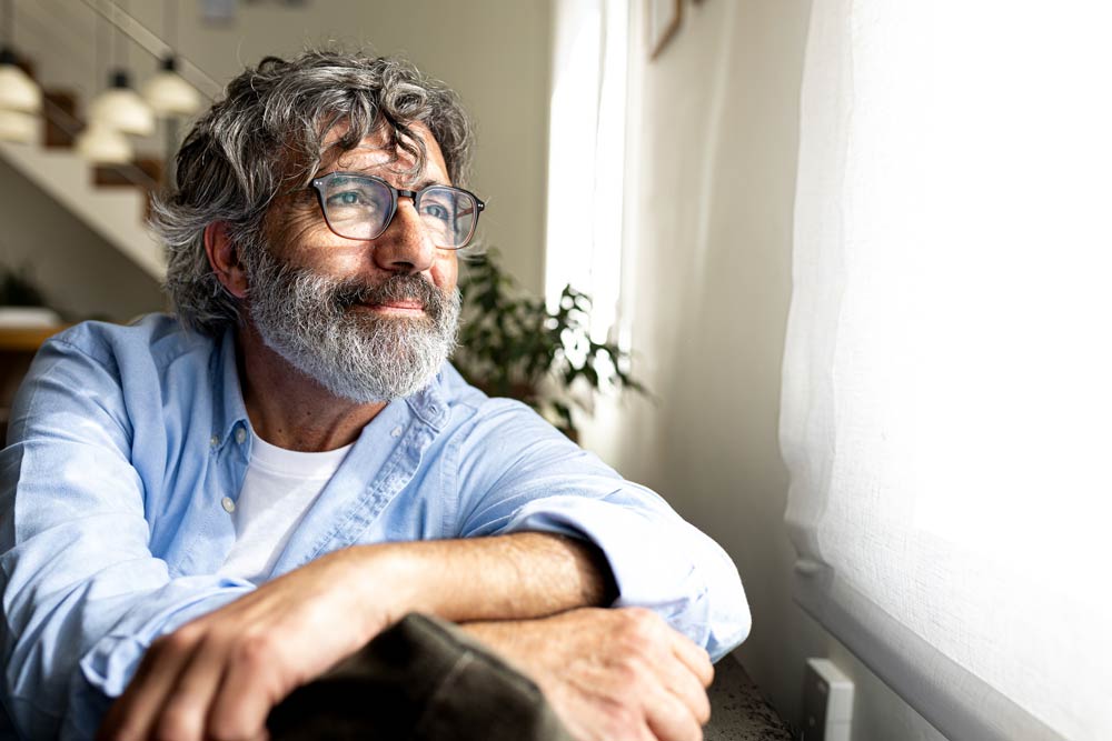 Older man looking out of a bright window with a thoughtful expression, symbolizing reflection on daily life challenges with Combined ADHD.